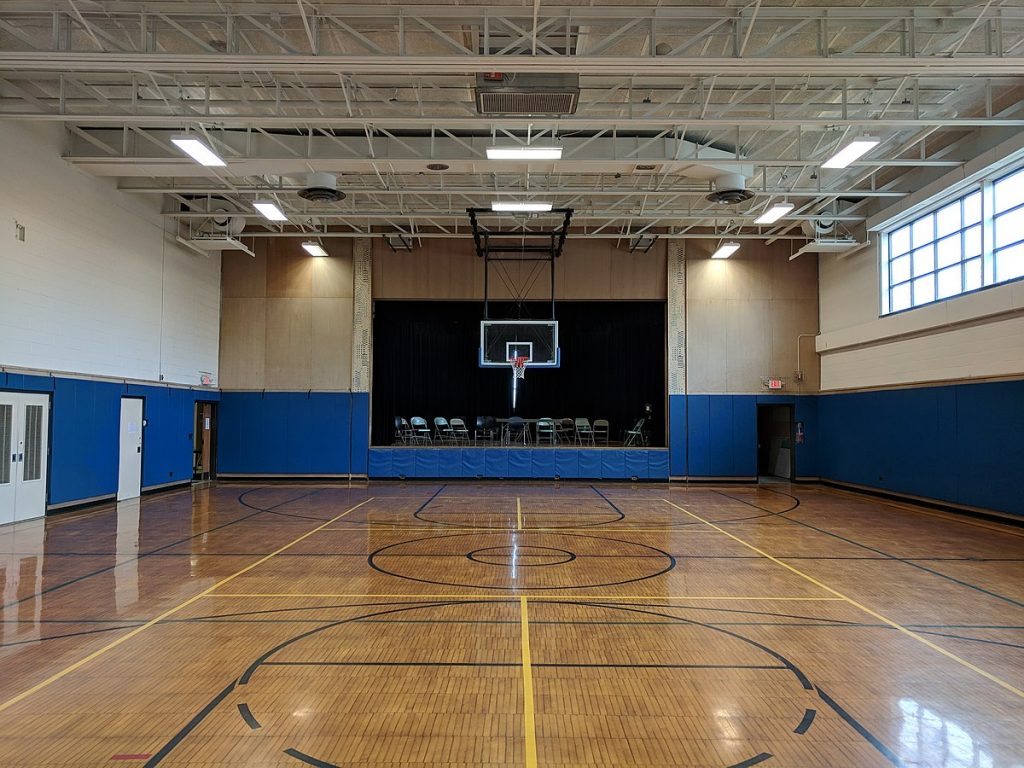 An empty school gymnasium with a basketball hoop suspended in front of a stage.