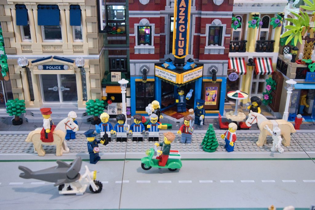 Street scene depicting sets from the Lego Modular Buildings series including a police station and jazz club. Various figures are present in front, including horses, police officers, a shark on a motorcycle, and a barbershop quartet.